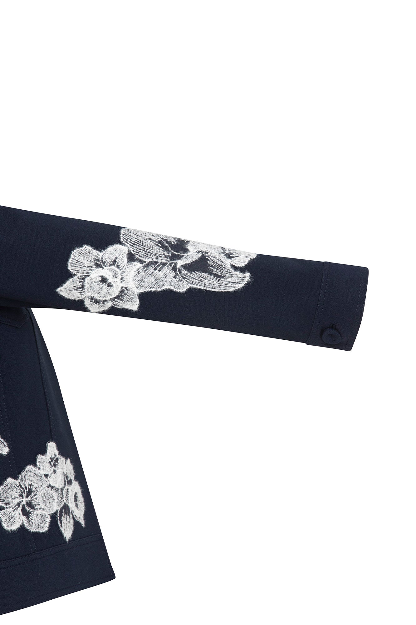 The Chaqueta Jean Flor Hilo Navy — sleeve close-up showing large-scale white iris and peony botanical embroidery in dense white thread with fine etched petal detail running from mid-arm to cuff on structured navy crepe, with single navy button closure at wrist