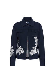 The Chaqueta Jean Flor Hilo Navy — full front view showing cropped structured navy crepe jacket with spread collar, concealed closure, two chest flap pockets, and large-scale white botanical iris and peony embroidery on lower front panels and sleeves