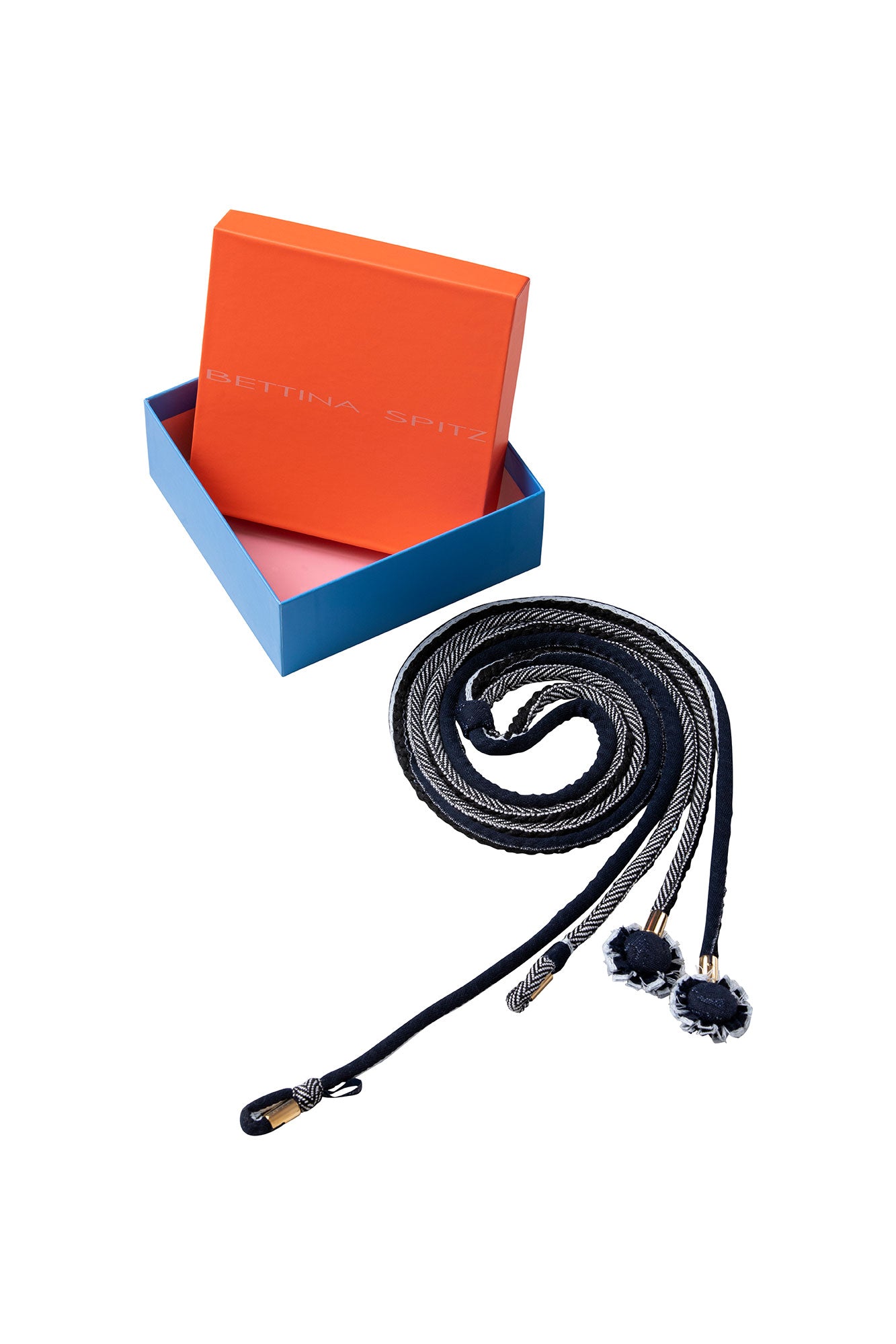The Cordón Doble — coiled beside the Bettina Spitz signature gift box with orange lid and blue base, showing both herringbone braid cords and black fabric pompom rosette ends