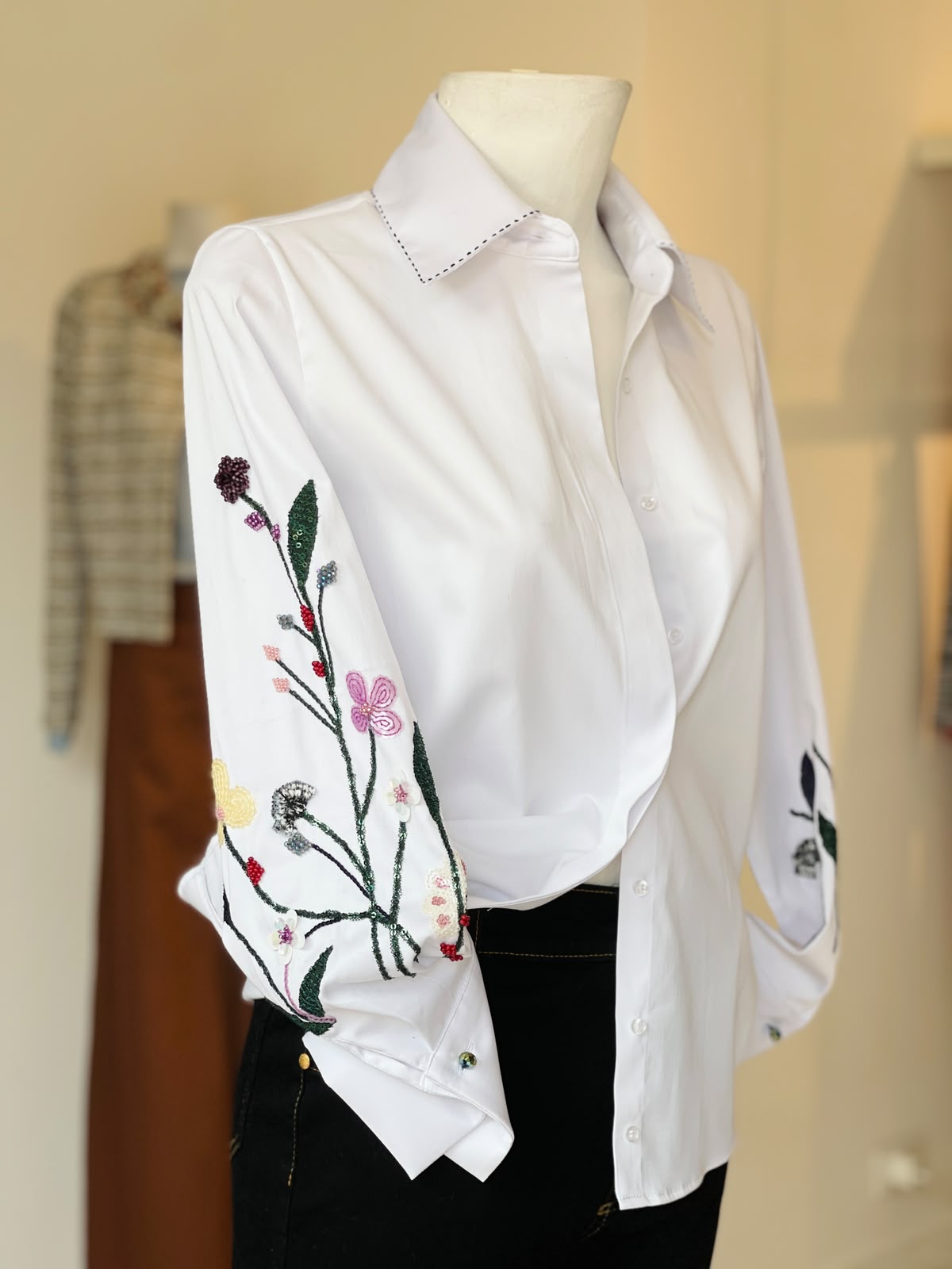 The Sol Flor White Shirt — styled on mannequin showing embroidered wildflower sleeves with purple berries, pink butterfly, yellow blooms and green stems