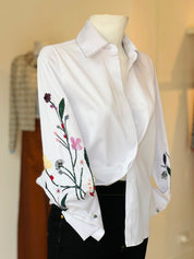 The Sol Flor White Shirt — styled on mannequin showing embroidered wildflower sleeves with purple berries, pink butterfly, yellow blooms and green stems